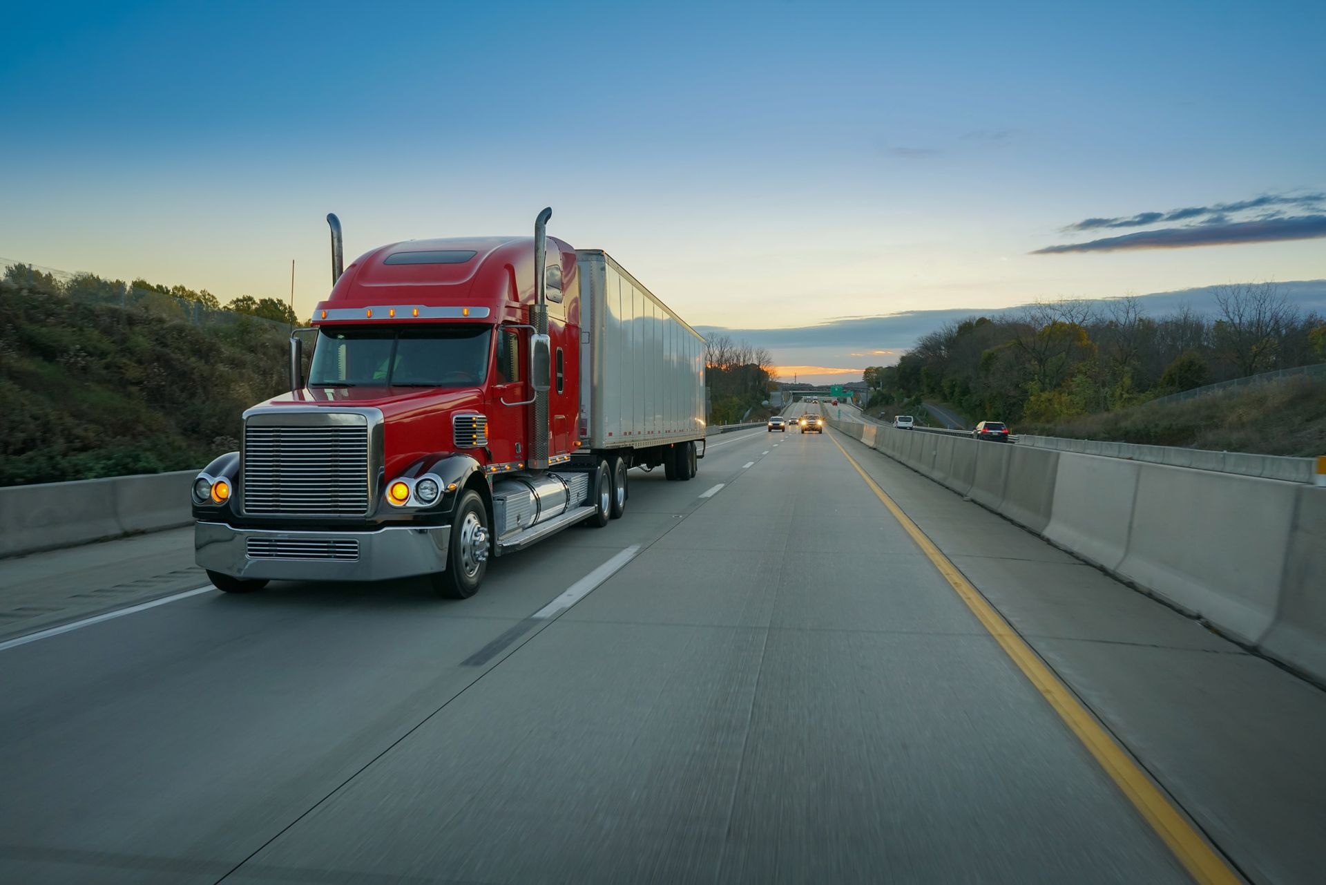 A red semi truck is driving down a highway at sunset.