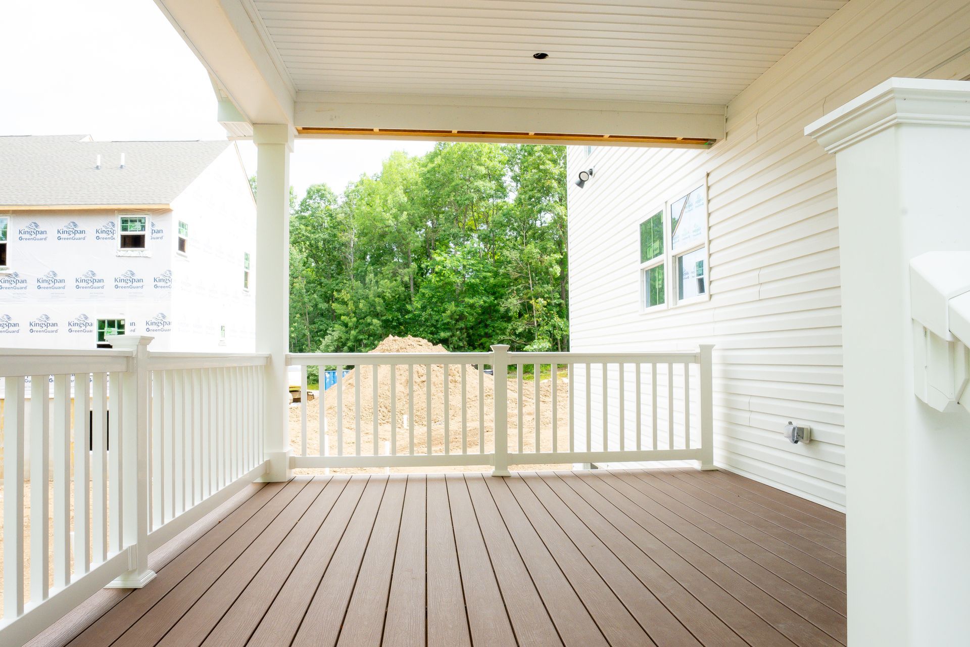Covered porch with white railing, brown decking, overlooking yard with trees, and a white building under construction.