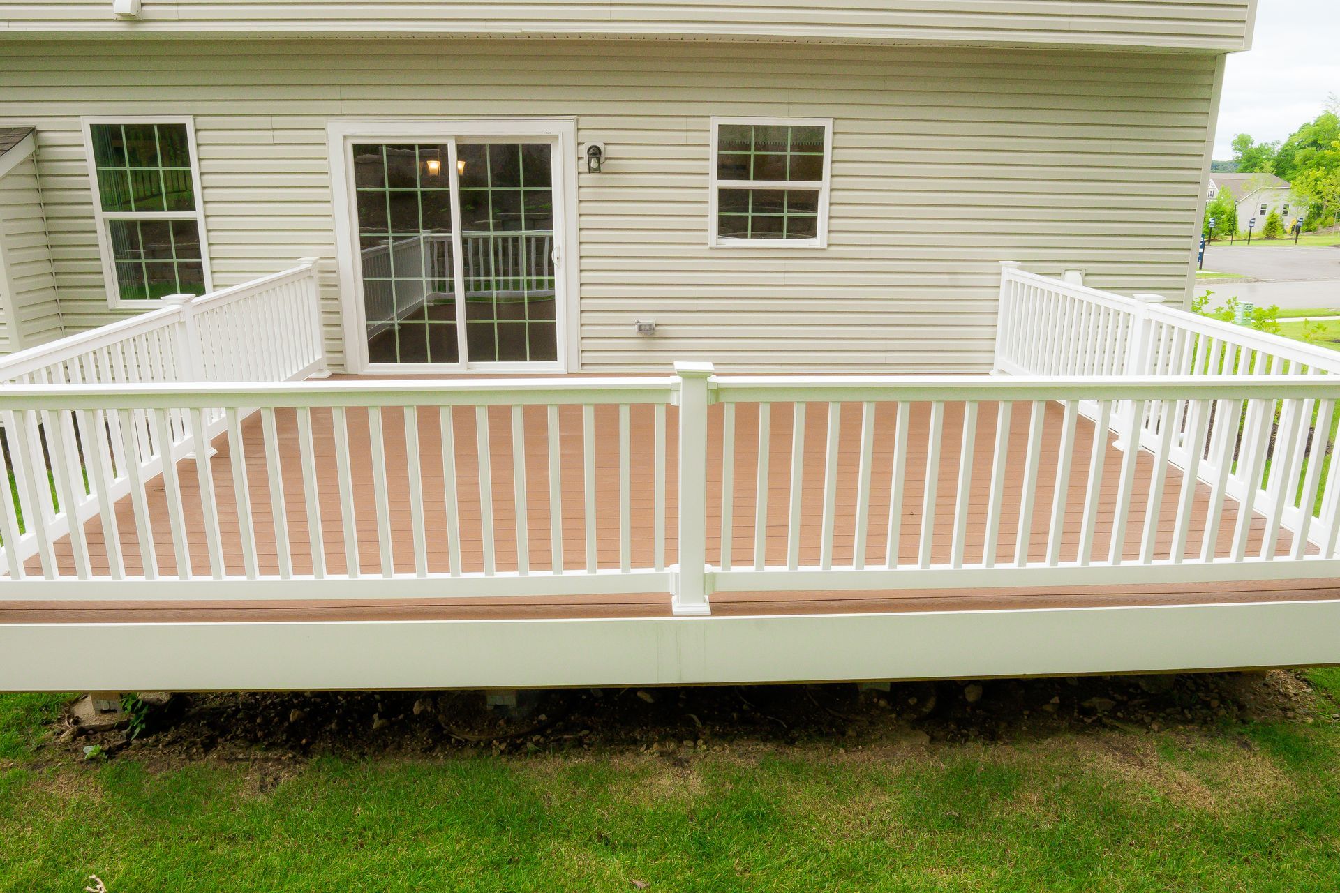 White railing surrounds a brown wooden deck attached to a light beige house with a sliding glass door.