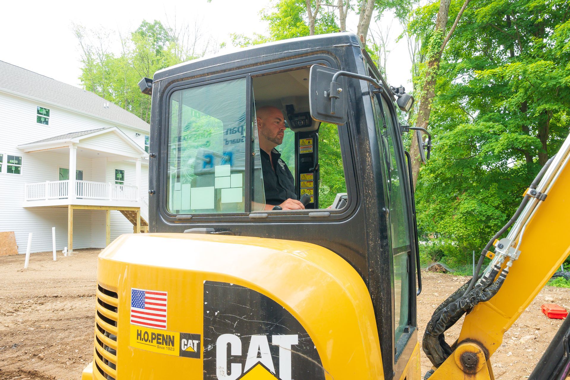 Man in a yellow CAT excavator cab, operating it outside a white building. American flag sticker on the machine.