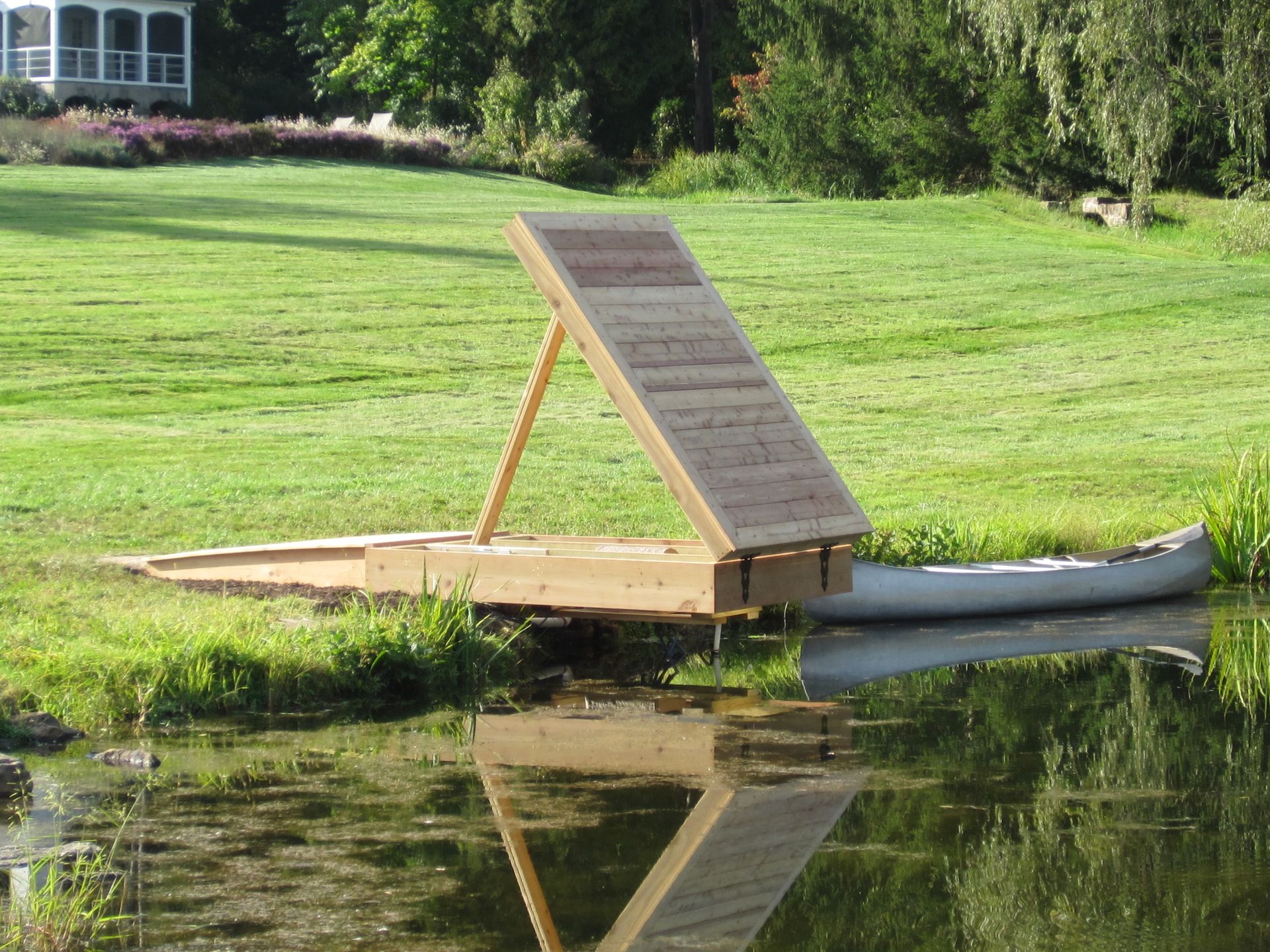 A wooden platform with a slanted ramp sits next to a canoe on a pond's edge.