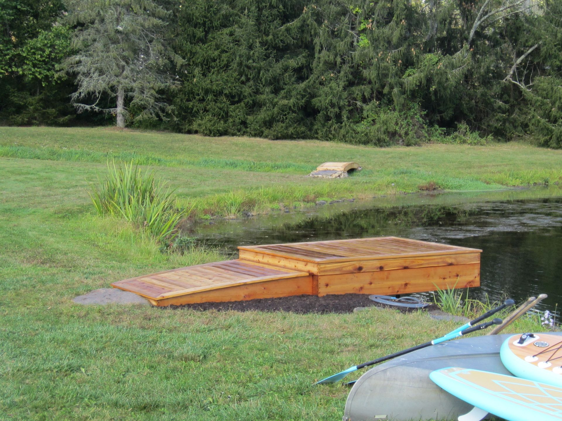 Wooden dock and ramp on pond's edge, built into the grass.  Paddleboards and poles visible. Green landscape.