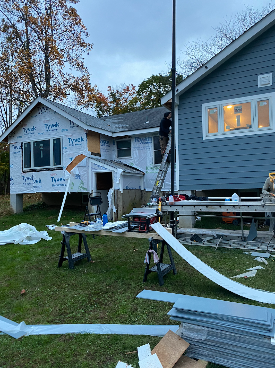 Construction on a house: worker on a ladder, siding installation, blue, white, gray, tools scattered on the ground.