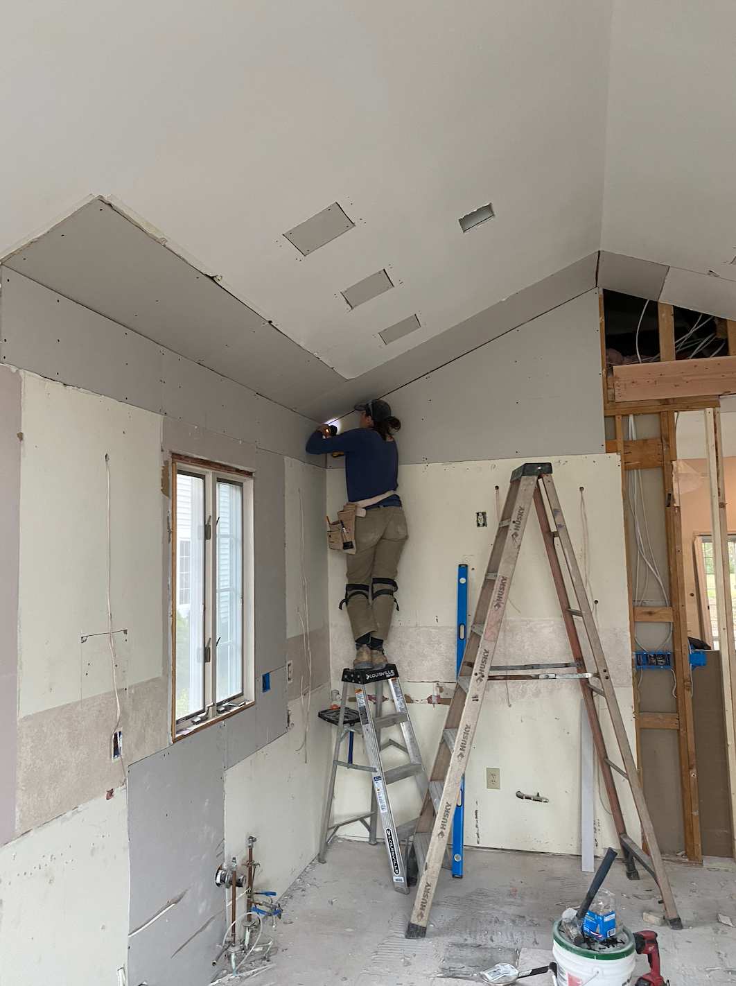 Person on a stepladder hanging drywall in a room under construction. Drywall and framing are visible.