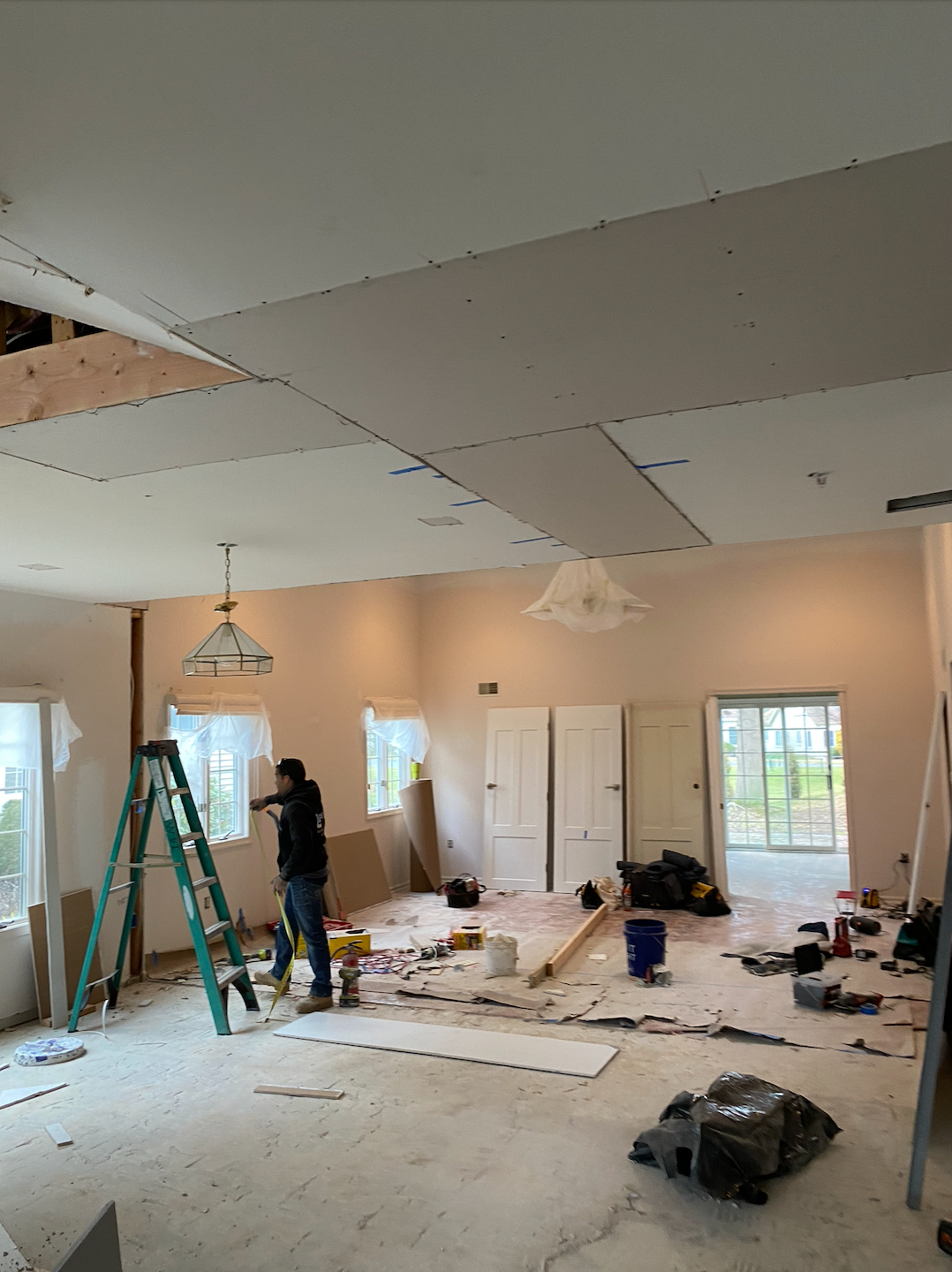 Man installing drywall on a ceiling in a room under renovation, tools and materials scattered on the floor.