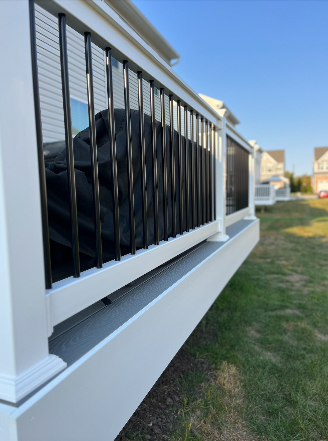 White deck with black railings, overlooking a green lawn and neighboring houses under a blue sky.