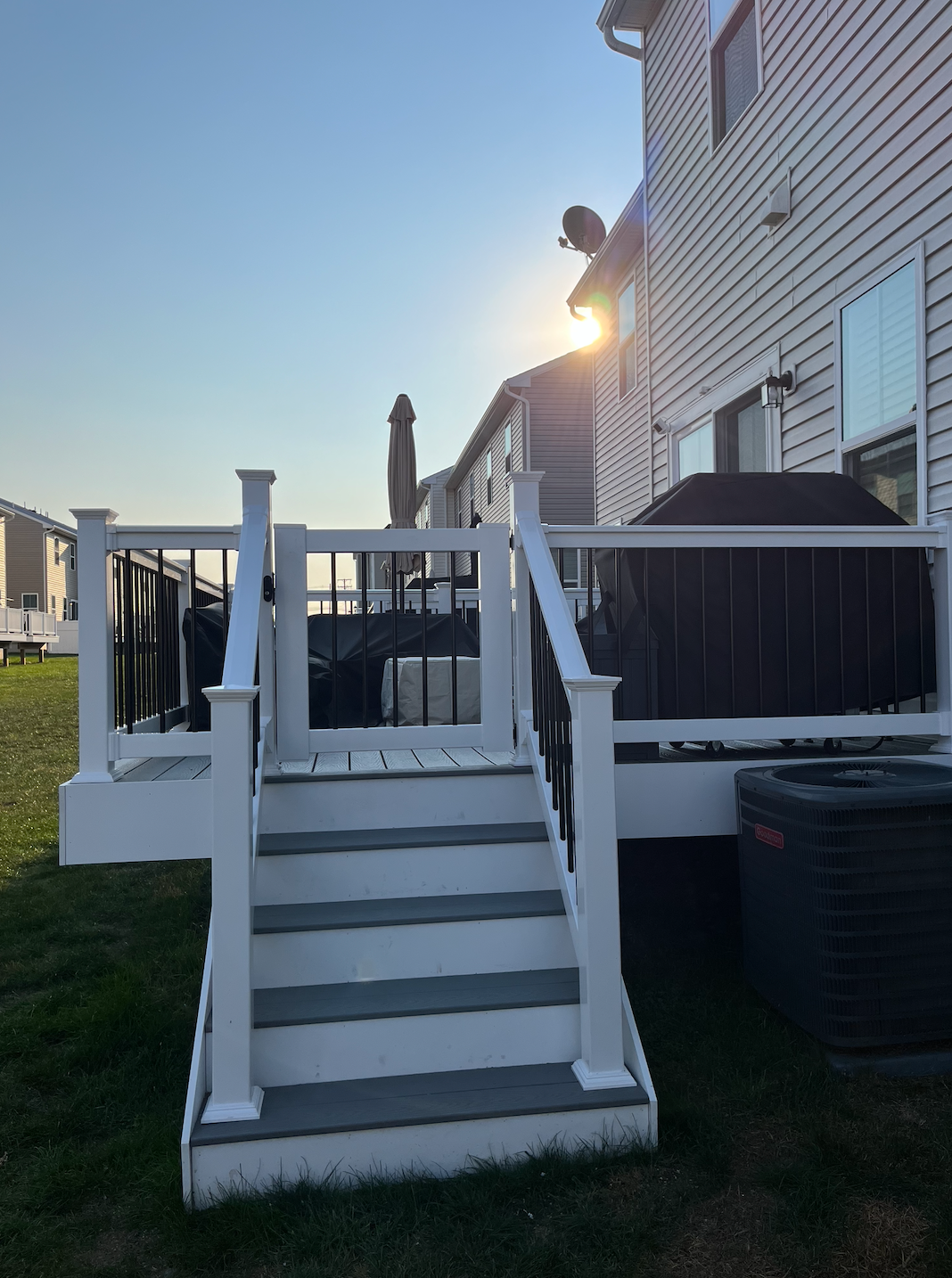 A white deck with steps leading down to a grassy yard, the sun shining on a house.