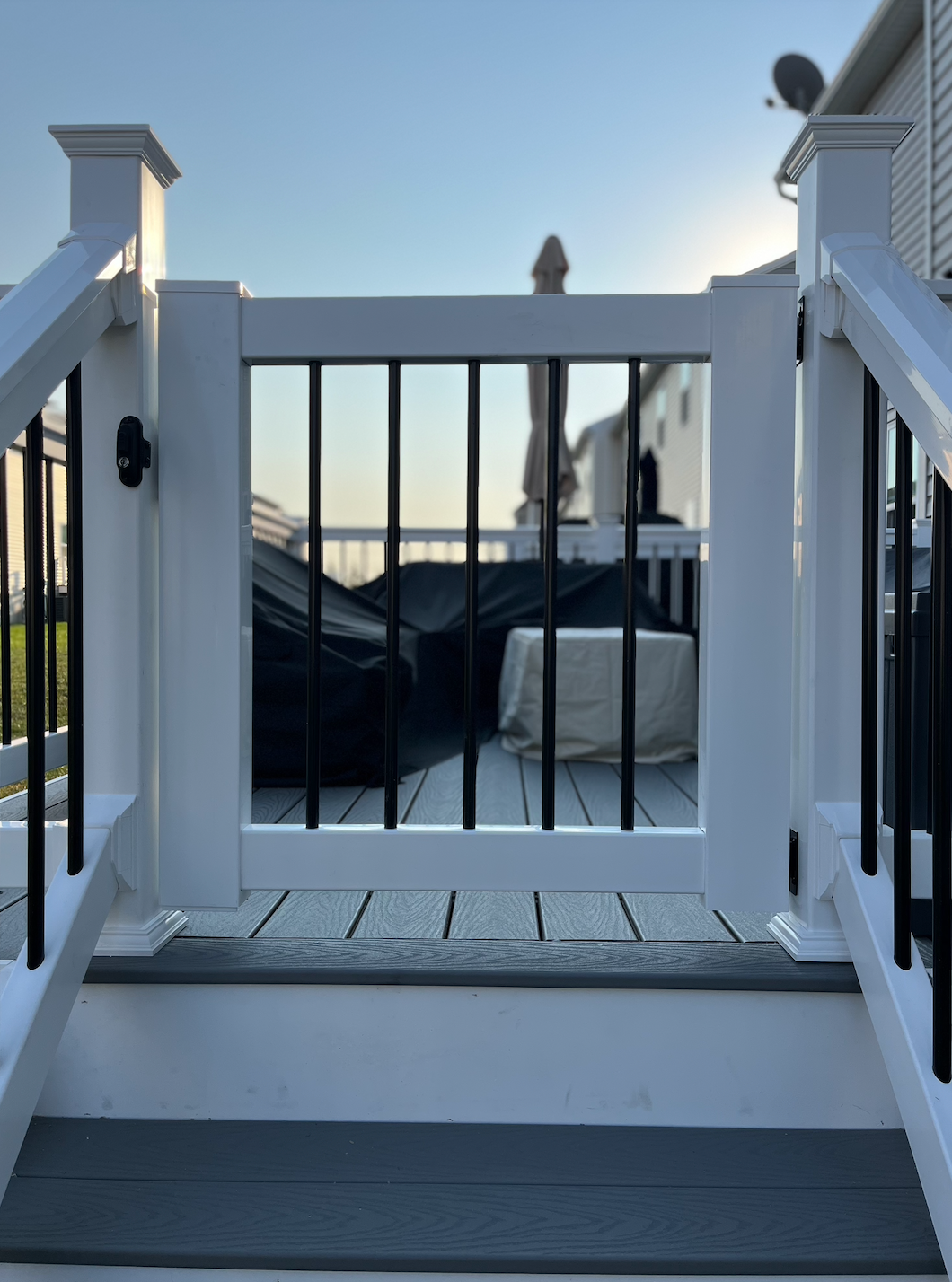 White deck gate with black vertical bars, leading to a back deck, sunny day.