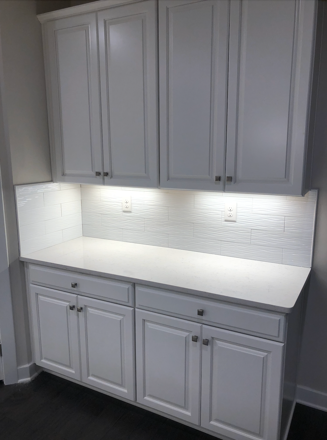 White kitchen cabinets with countertop and backsplash, illuminated under cabinet lighting.