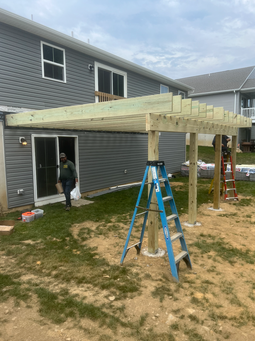 Construction of a wooden pergola attached to a gray house. A person walks near the door. Blue ladder in front.