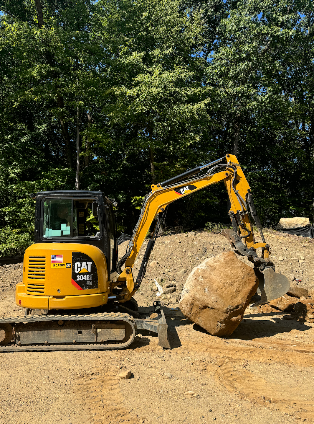 Yellow CAT excavator lifting a large rock in a gravel area; trees in the background.