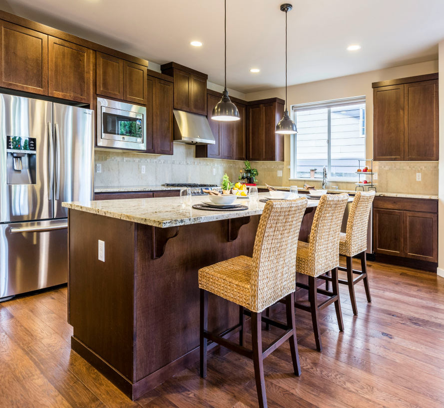 Modern white kitchen with island, stovetop, and silver vent hood. Bar stools, cabinetry, and black countertop.