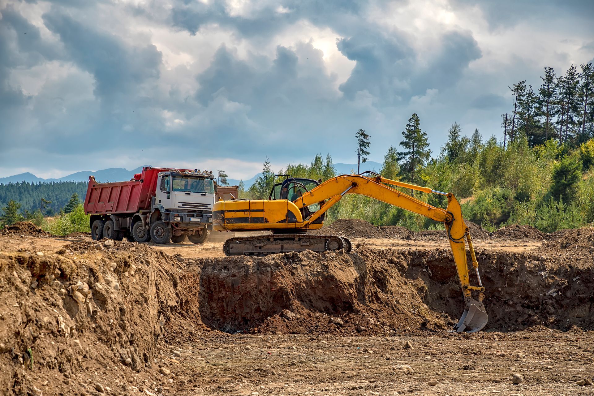 A yellow excavator digs dirt, loading a red dump truck at a construction site under a cloudy sky.