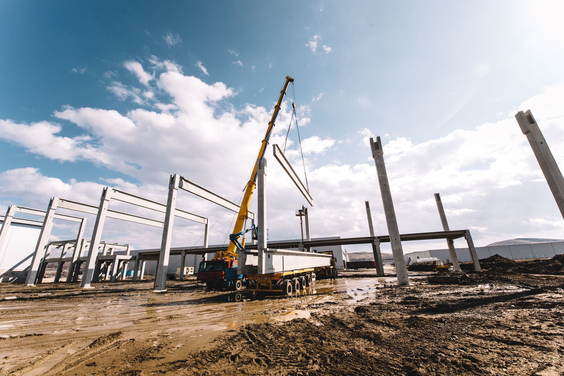 Construction site with crane lifting concrete beams; sunny, cloudy sky overhead.