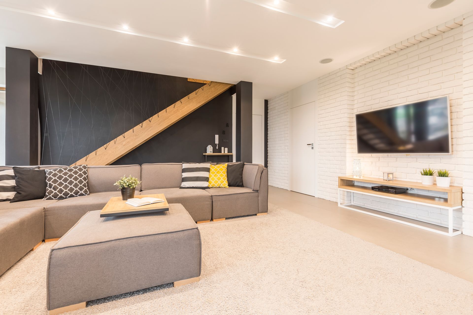 Modern living room with gray sectional sofa, patterned pillows, rug, TV on white brick wall.