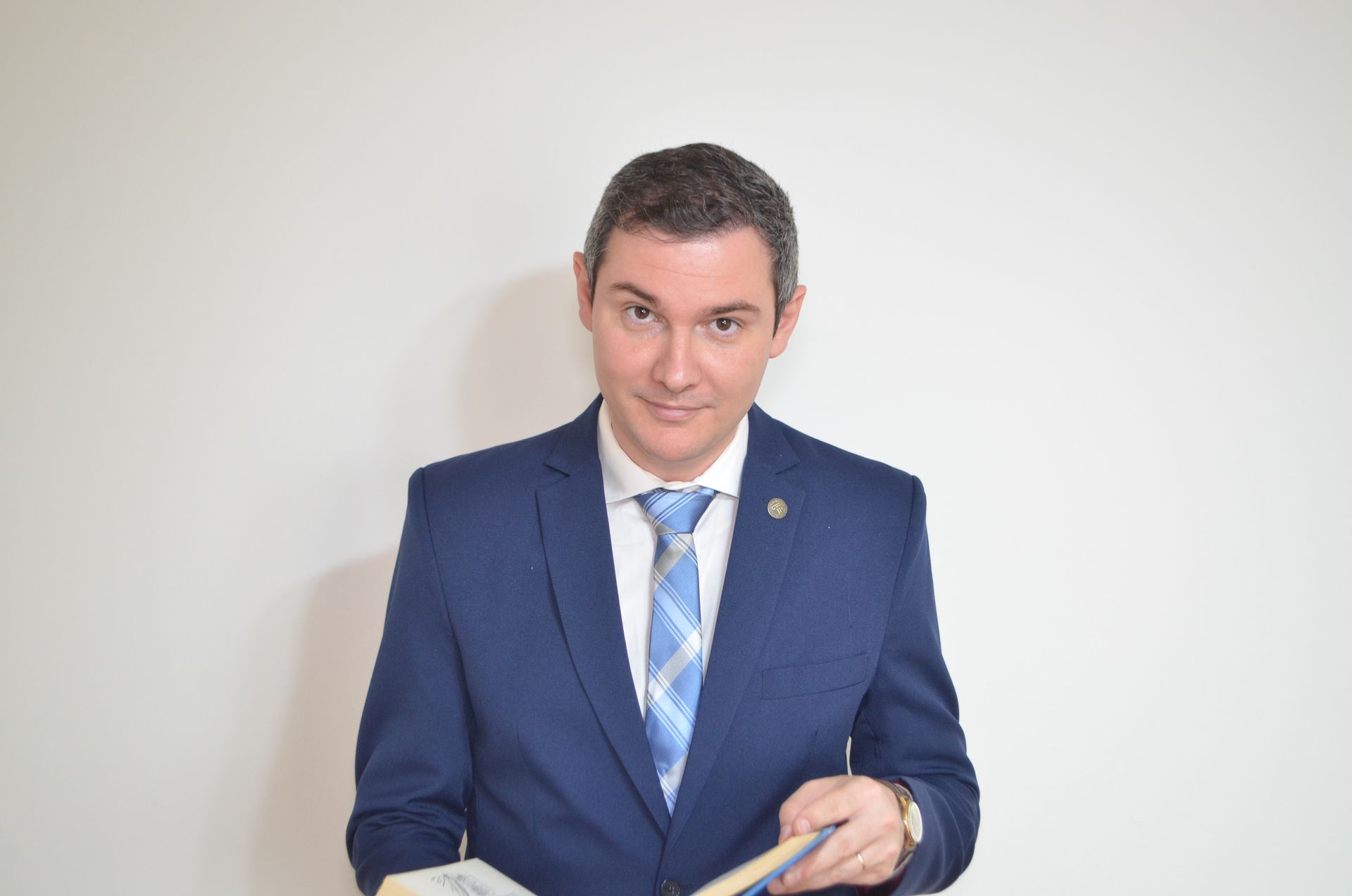 Hombre con traje azul, sonriente, sosteniendo un libro, de pie contra una pared blanca.