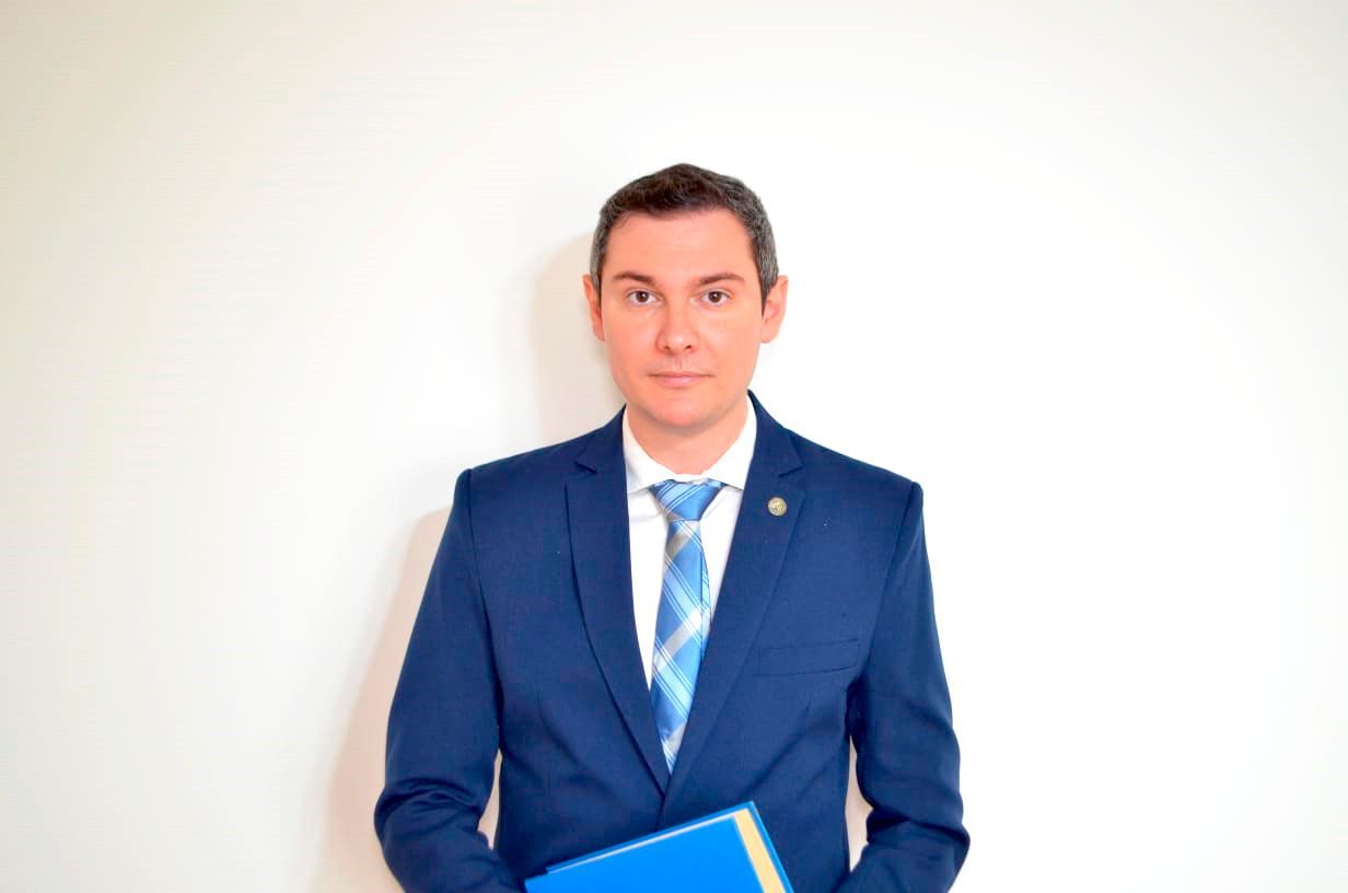 Hombre con traje azul, sosteniendo una carpeta azul, de pie frente a una pared blanca.