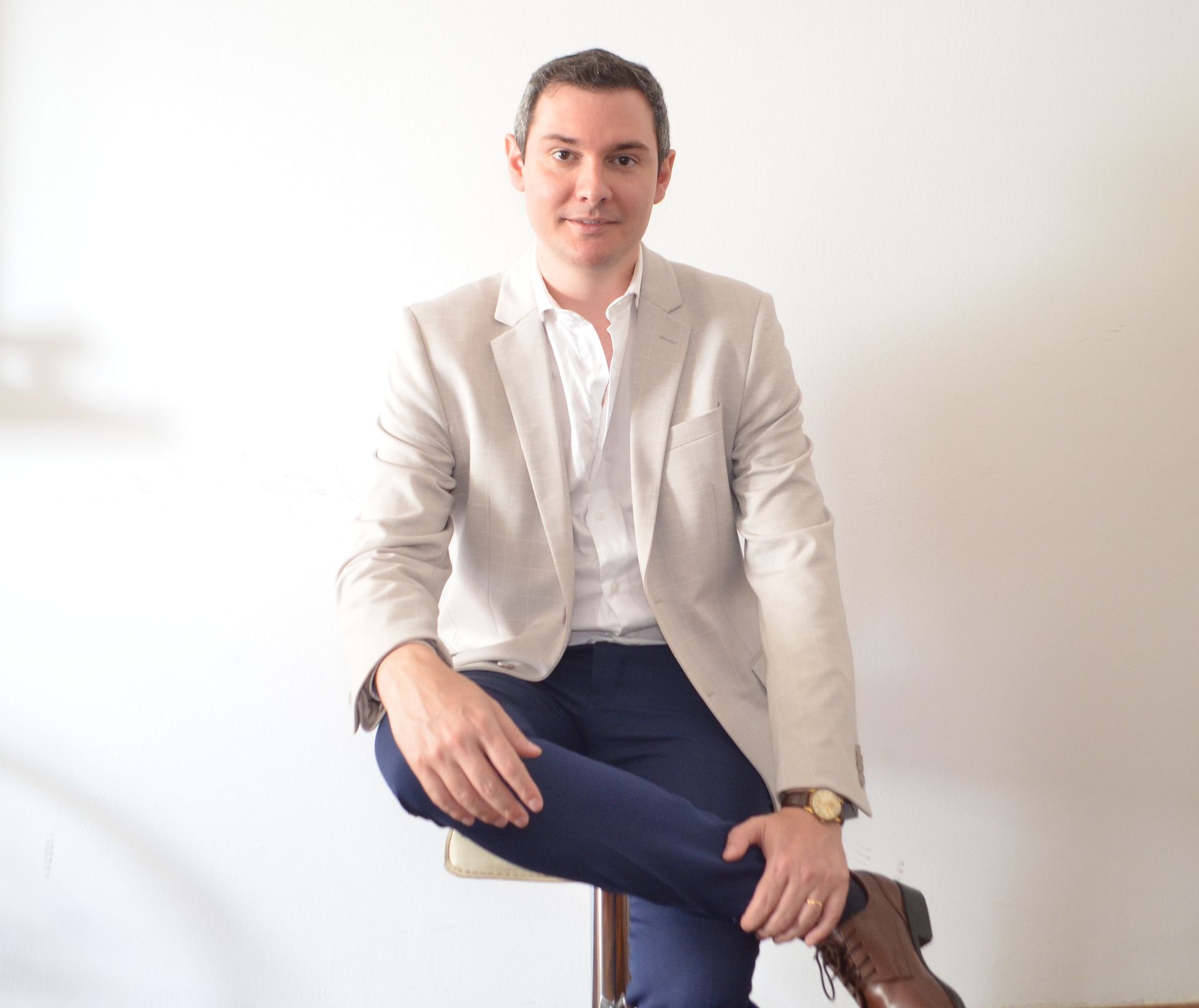 Hombre con chaqueta y pantalón, sentado en un taburete, mirando hacia el espectador, fondo de pared blanca.