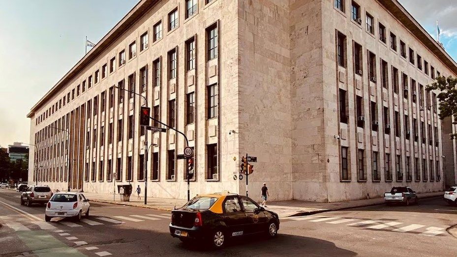 Edificio grande de color beige en una intersección urbana con tráfico; hay algunos coches y un peatón.