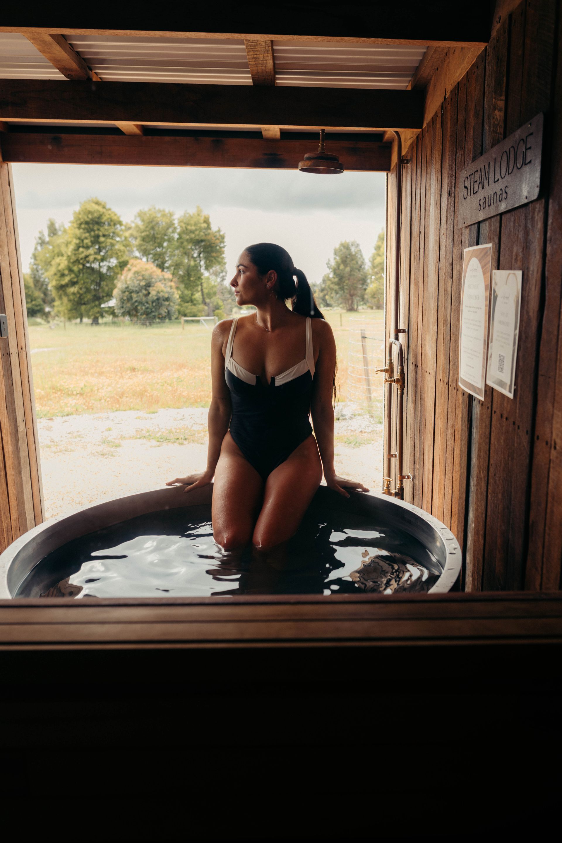 Woman in black swimsuit in outdoor wooden tub, looking away, countryside in background.