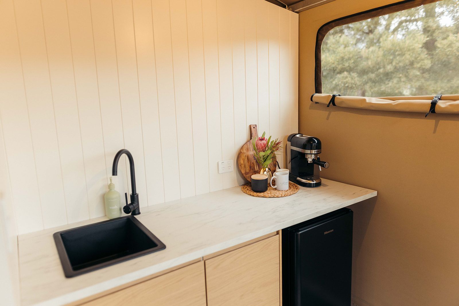 Kitchenette with black sink and faucet, coffee maker, and mini-fridge. White countertop and walls, natural light from window.
