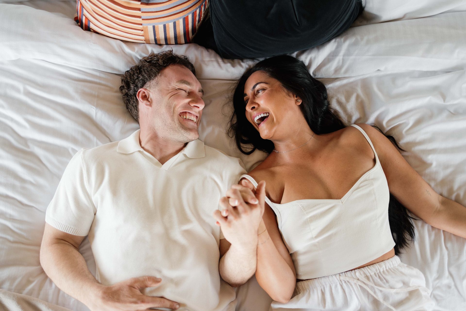 Couple in bed, laughing and holding hands, both wearing white clothing.
