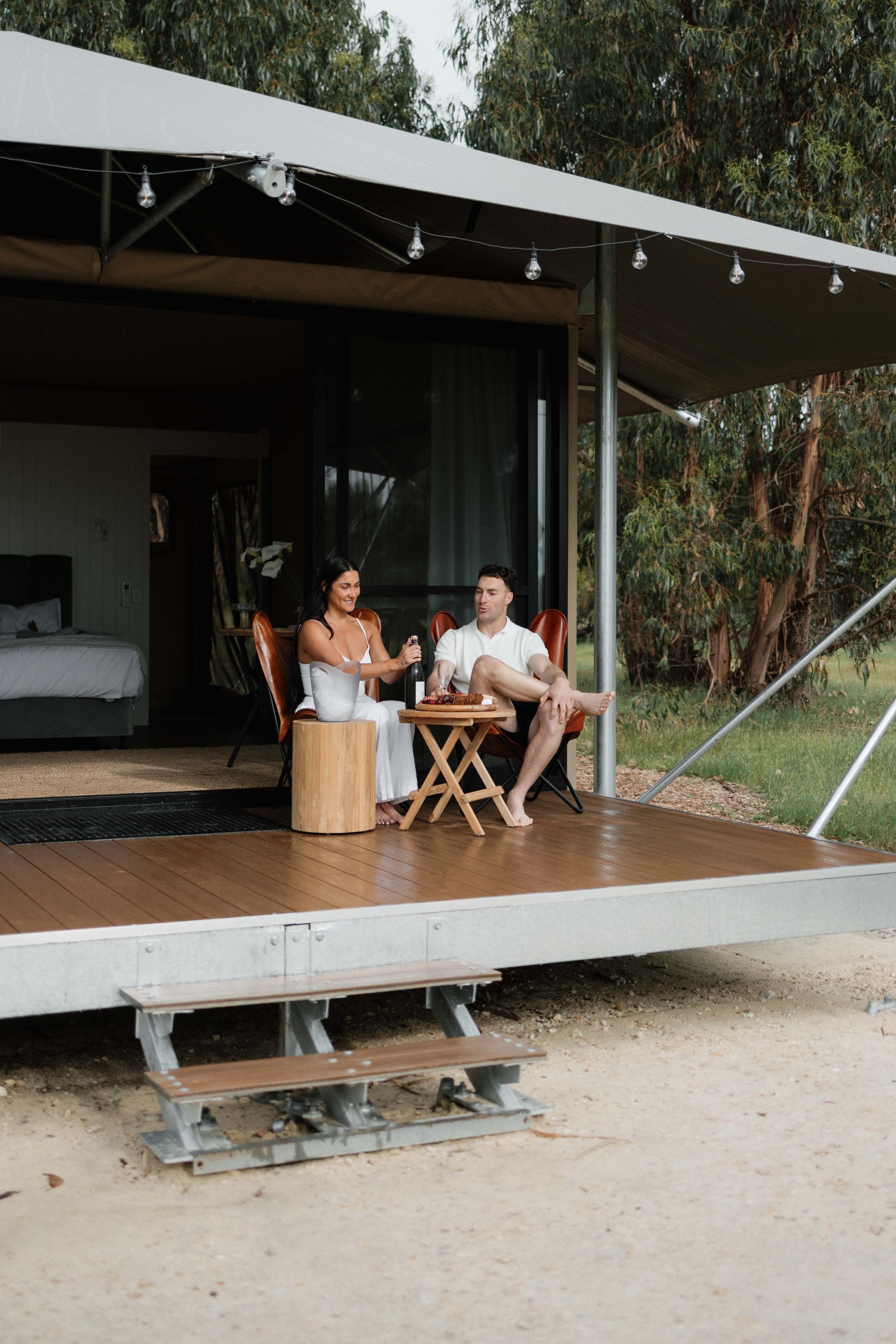 Couple sitting on a cabin porch, holding drinks, enjoying the outdoors.