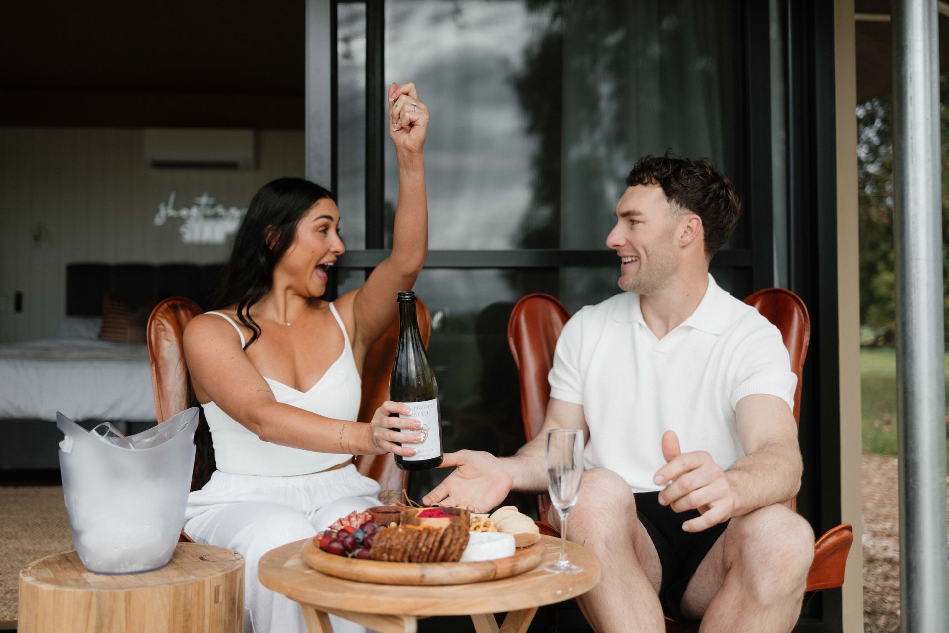Woman excitedly holding wine bottle with man, on patio with snacks and ice bucket.