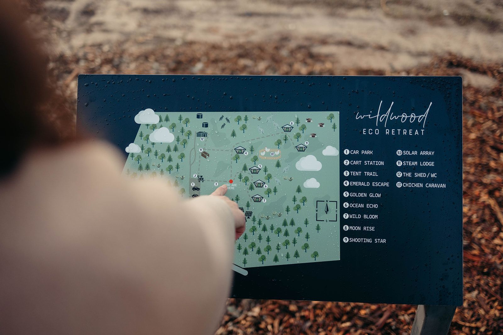 Person points to a map on a dark sign in an outdoor setting. The map has trees and buildings.