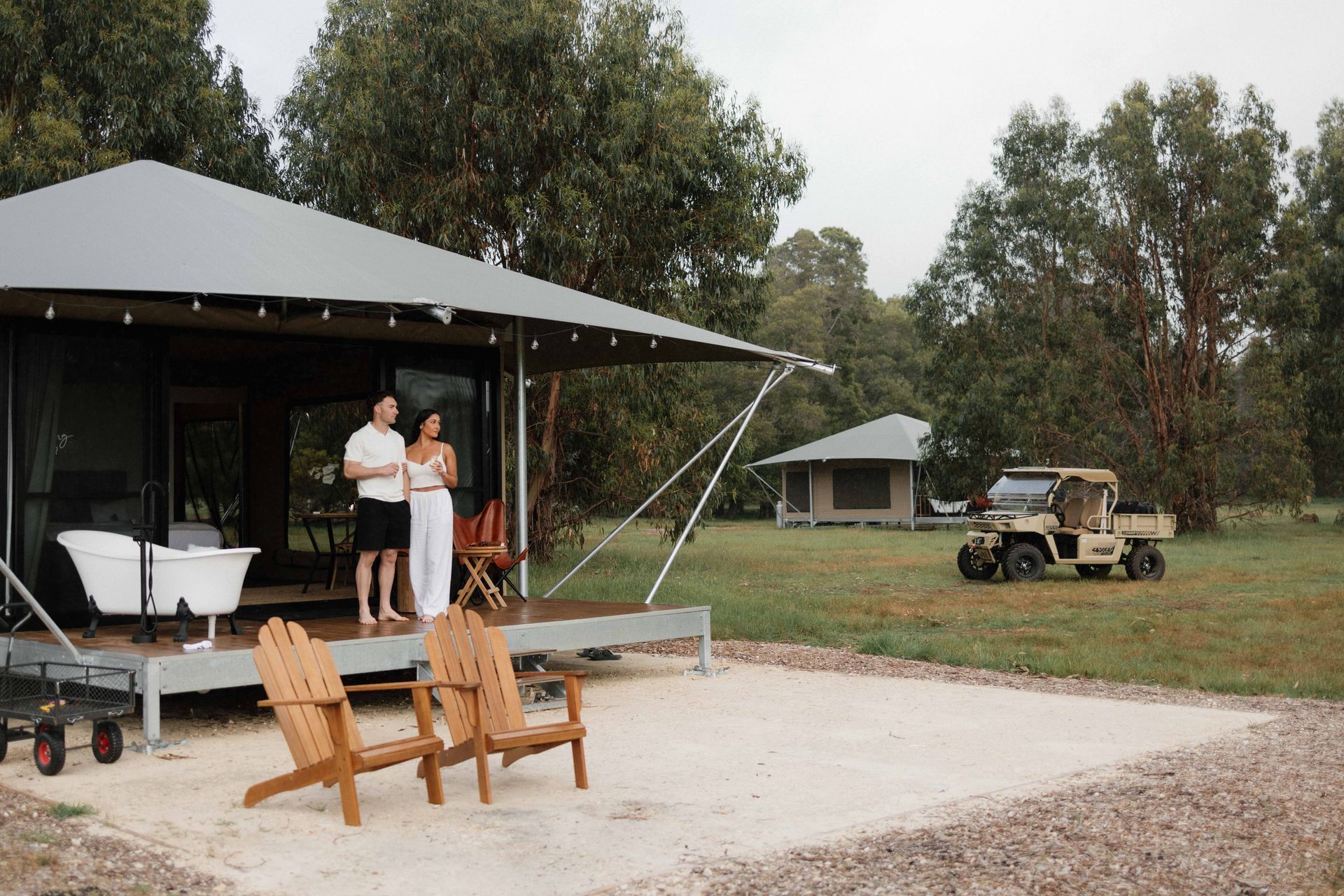 Couple on porch of cabin holding drinks, outdoor chairs, utility vehicle on the lawn.