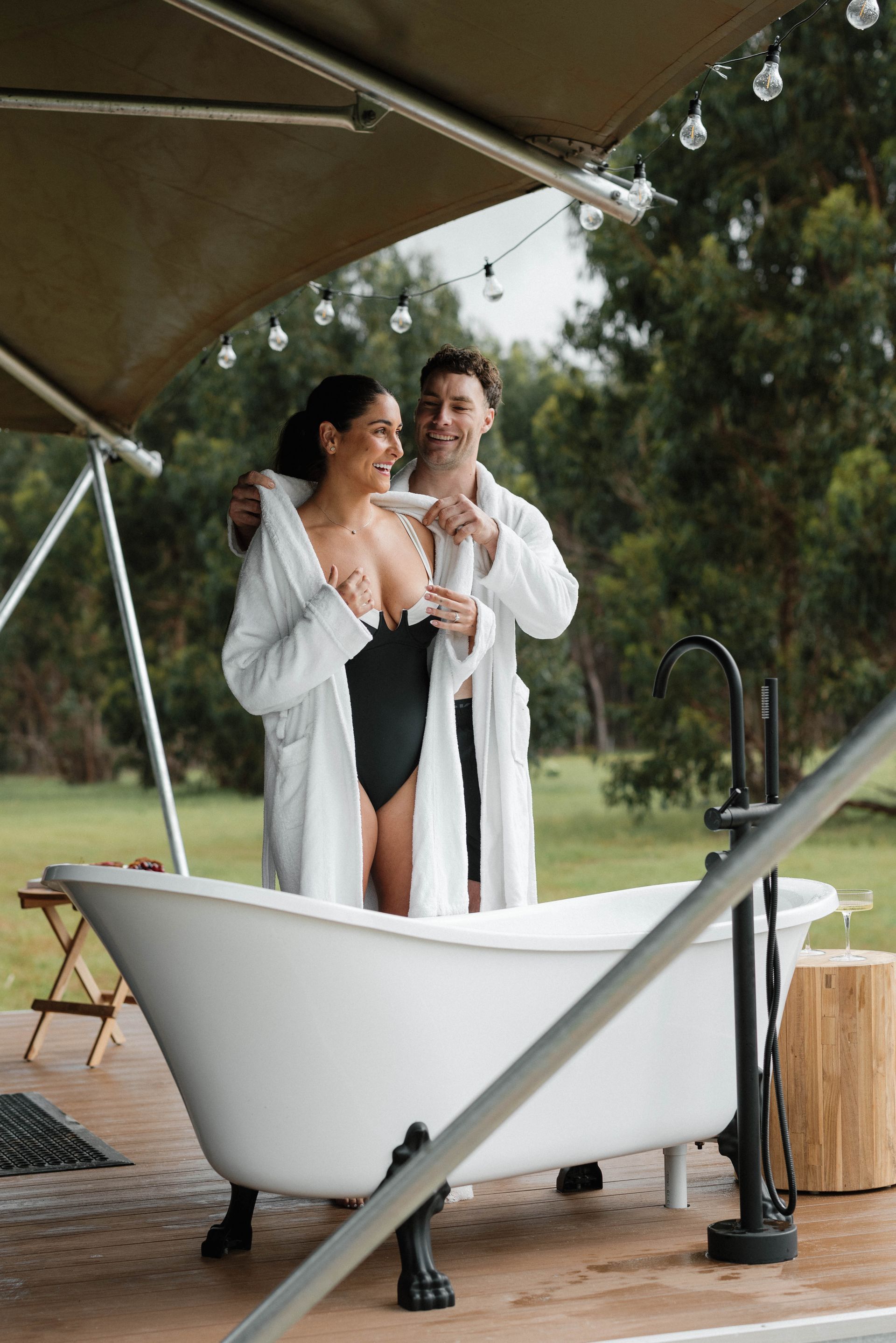 Couple in bathrobes, standing in a bathtub outdoors, smiling. Trees and tent in background.