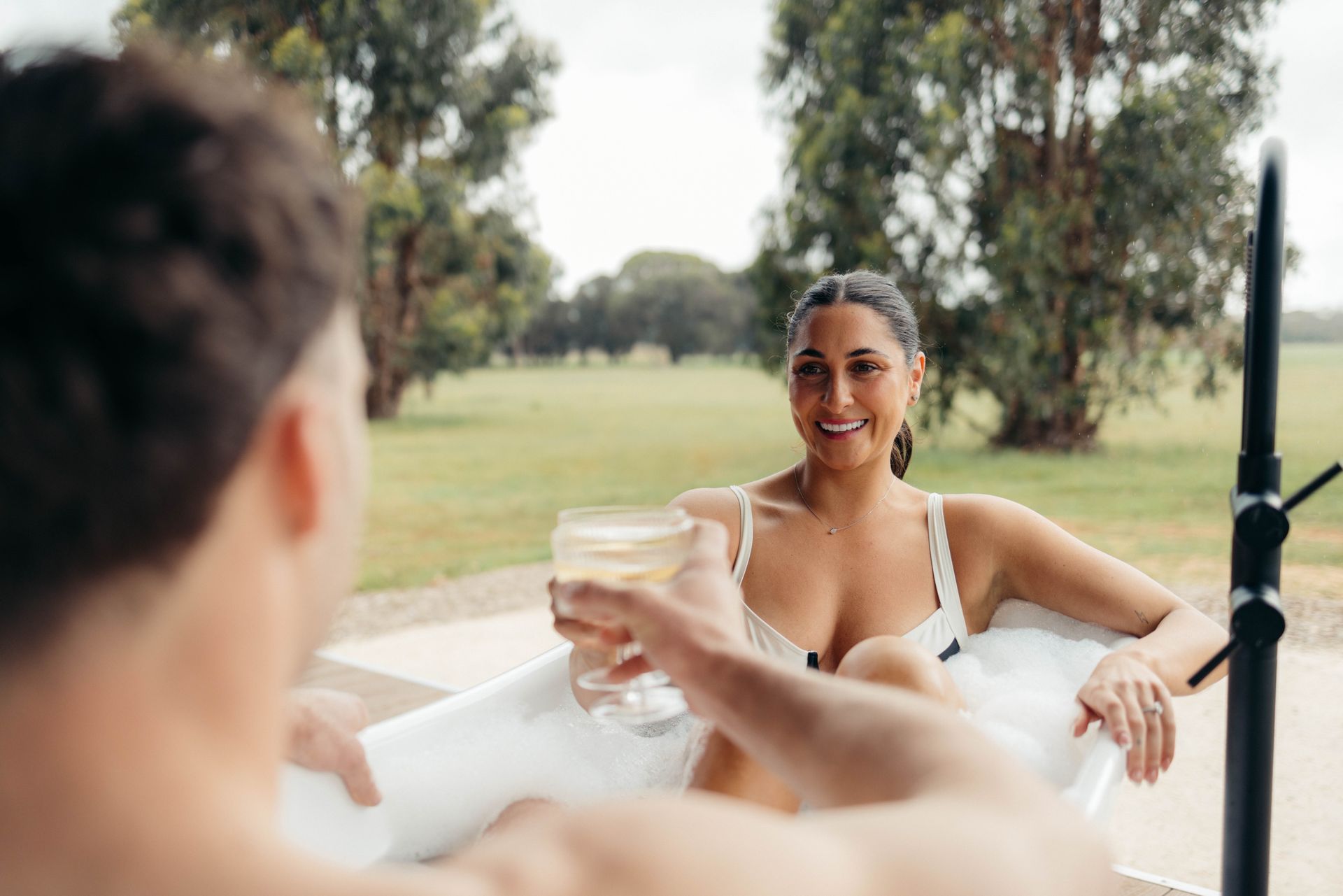 Woman in outdoor bathtub cheers with someone, smiling, holding a drink.