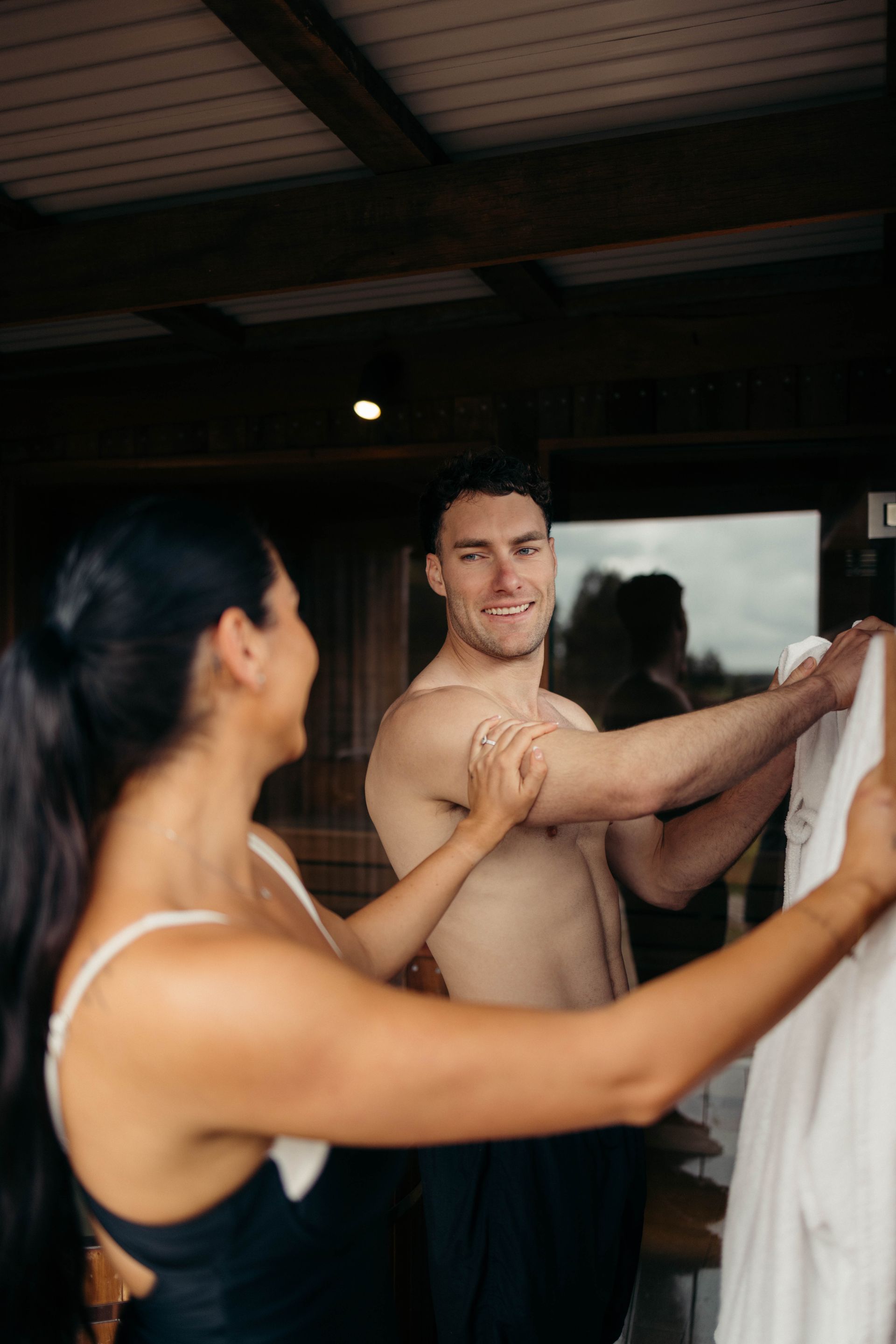 Woman helping a man with a white shirt; they're smiling inside a rustic building.