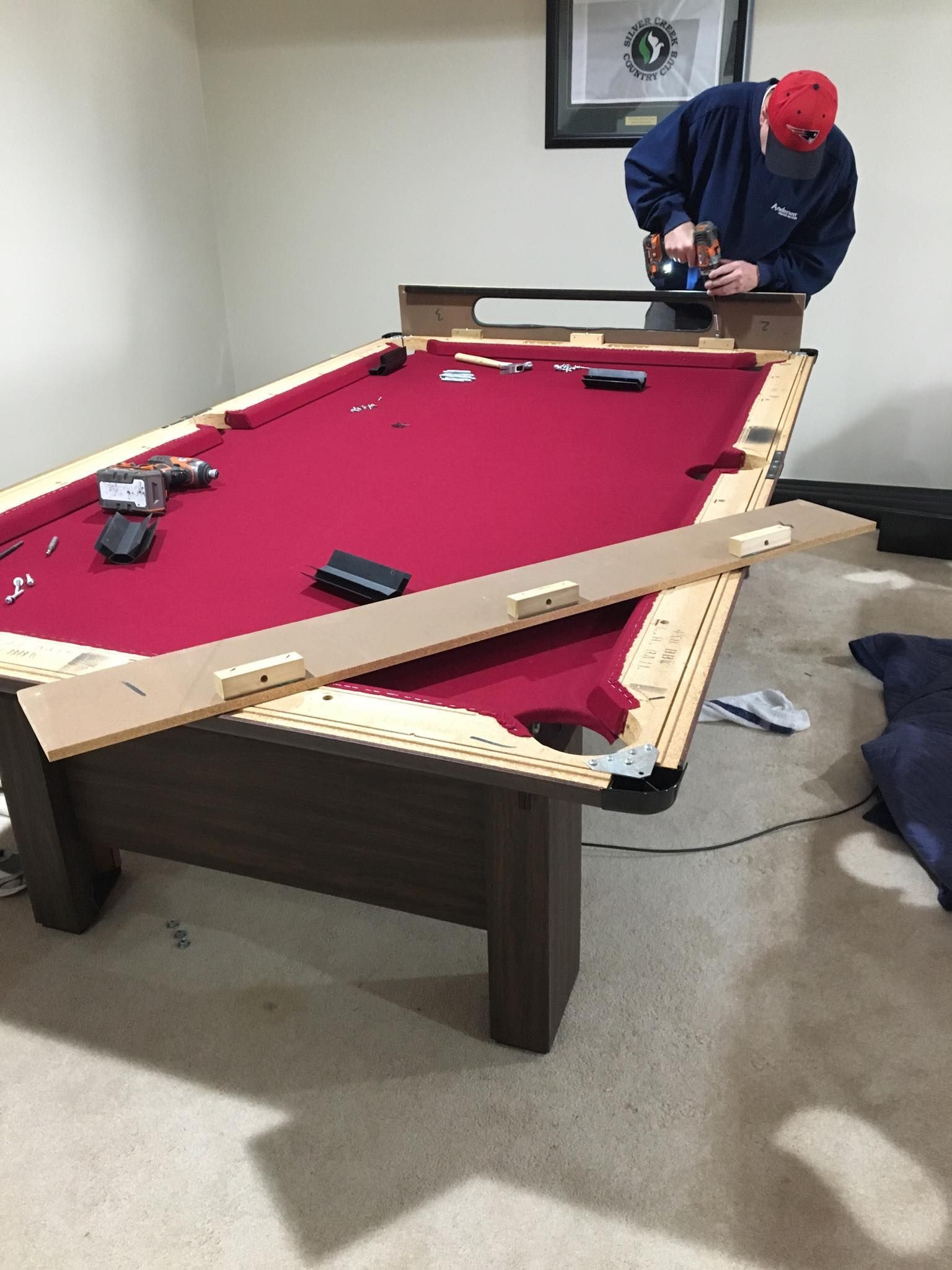 Man in red cap using a power drill on a pool table in a room. Man in red cap using a power drill on a pool table in a room.