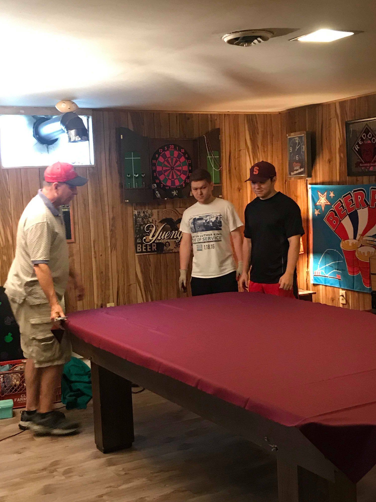 Three people covering a maroon pool table in a wood-paneled basement.