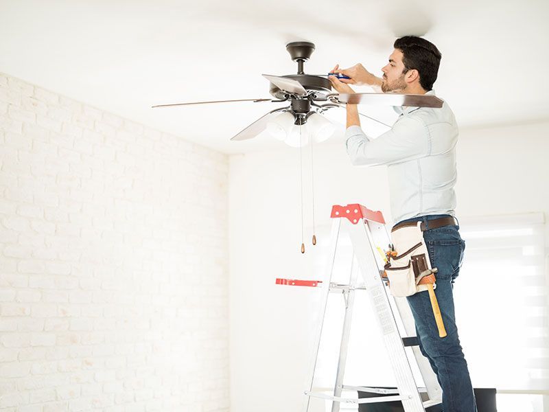 Man on a ladder installing a ceiling fan in a white room.