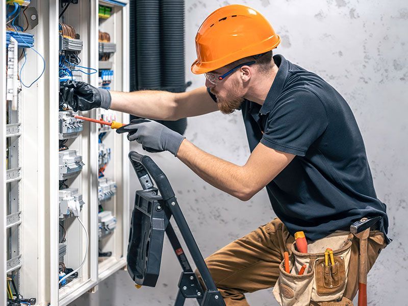 Electrician in orange hard hat, working on electrical panel, using screwdriver.