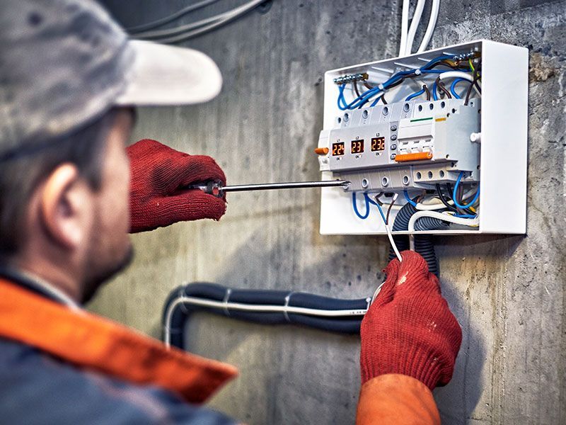 Electrician in red gloves working on a circuit breaker box, mounted on a concrete wall.