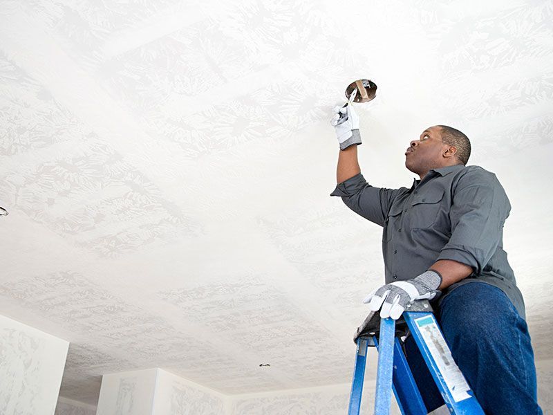 Man on ladder, inspecting an electrical fixture on a white textured ceiling, wearing work gloves.