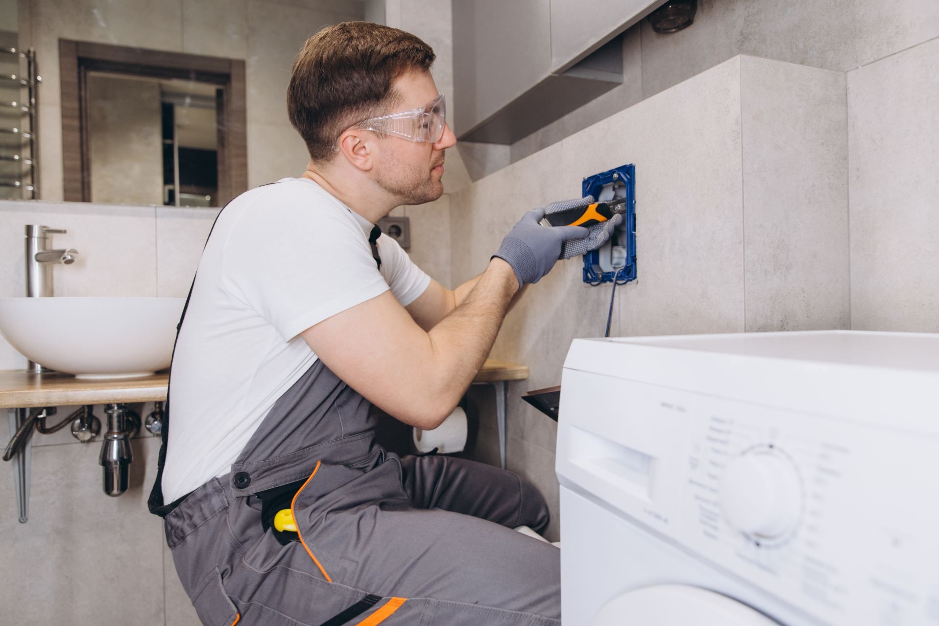 Electrician working on electrical outlet near a washing machine and sink, wearing safety glasses and gloves.