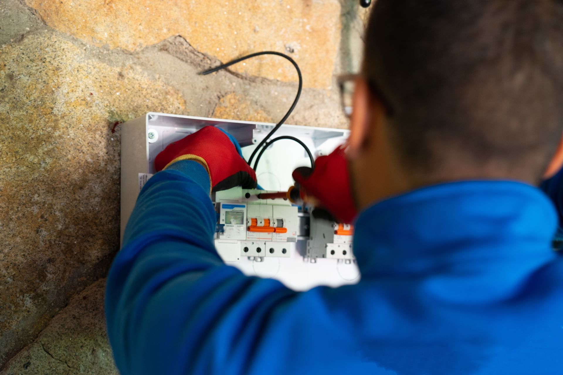 Electrician in red gloves works on circuit breaker box mounted to stone wall.