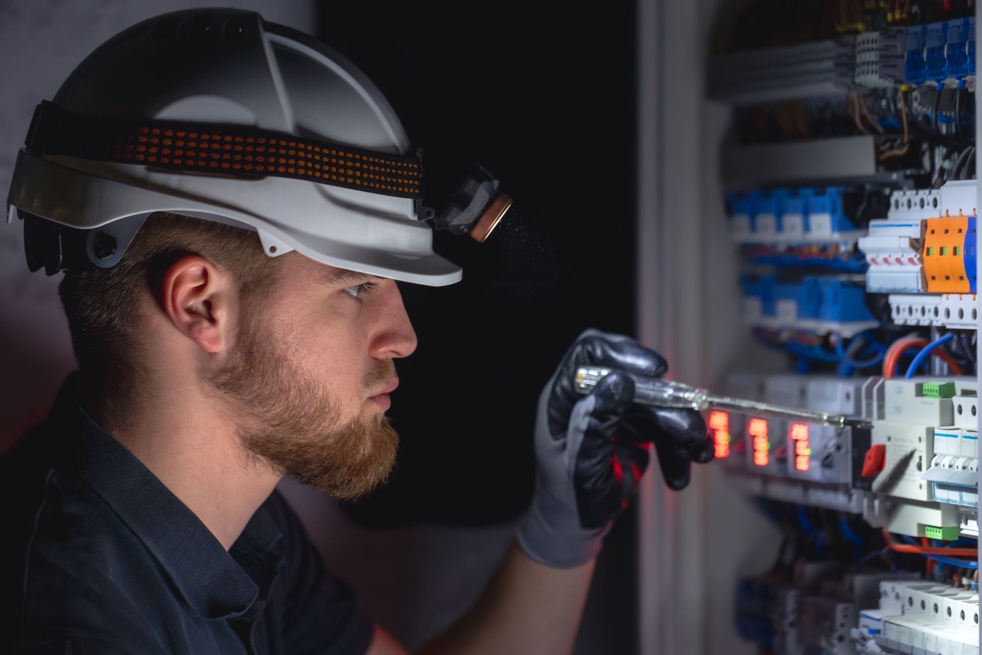 Electrician inspecting electrical panel wearing hardhat with headlamp.