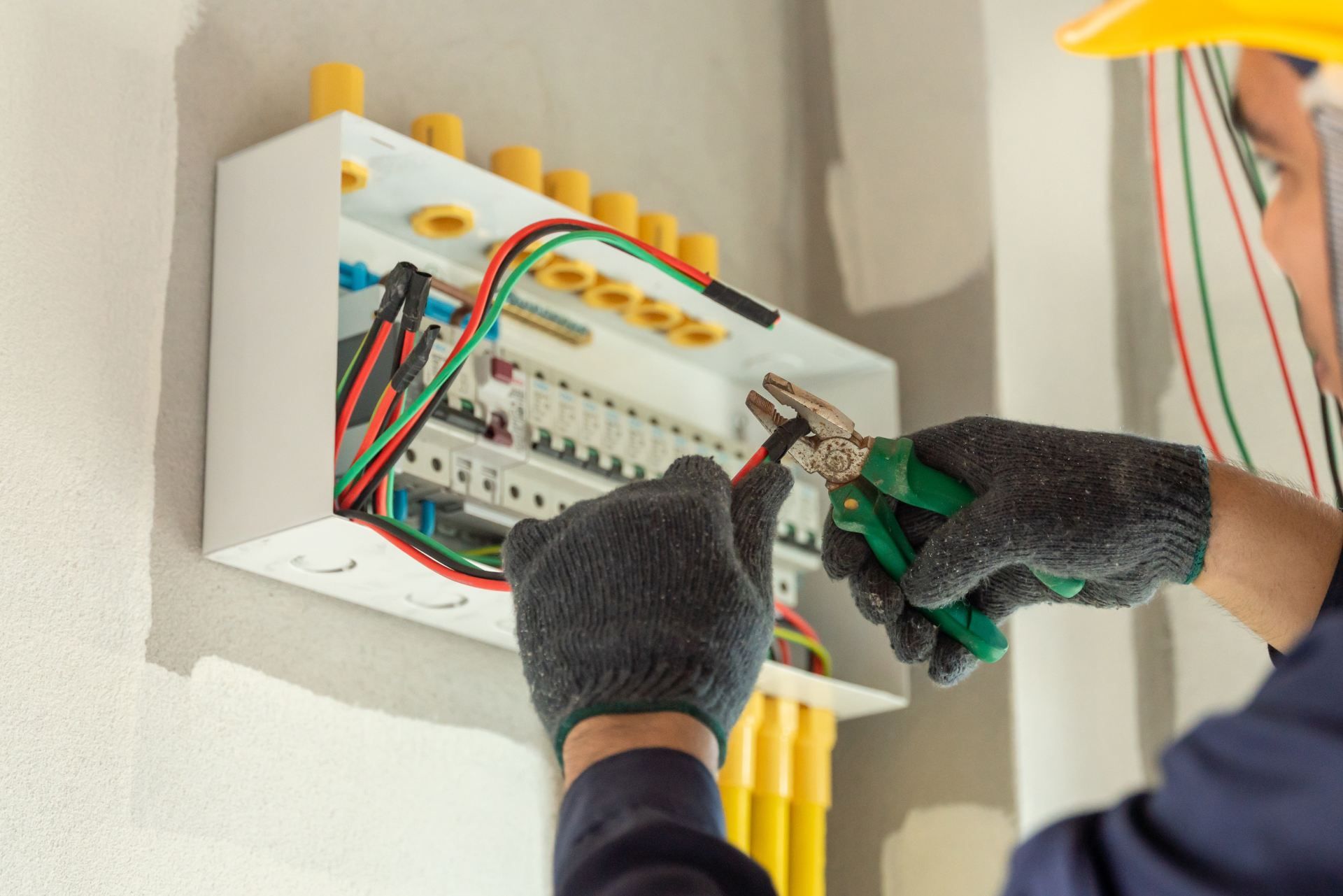 Electrician working on a circuit breaker box with pliers, wearing gloves and a hard hat.