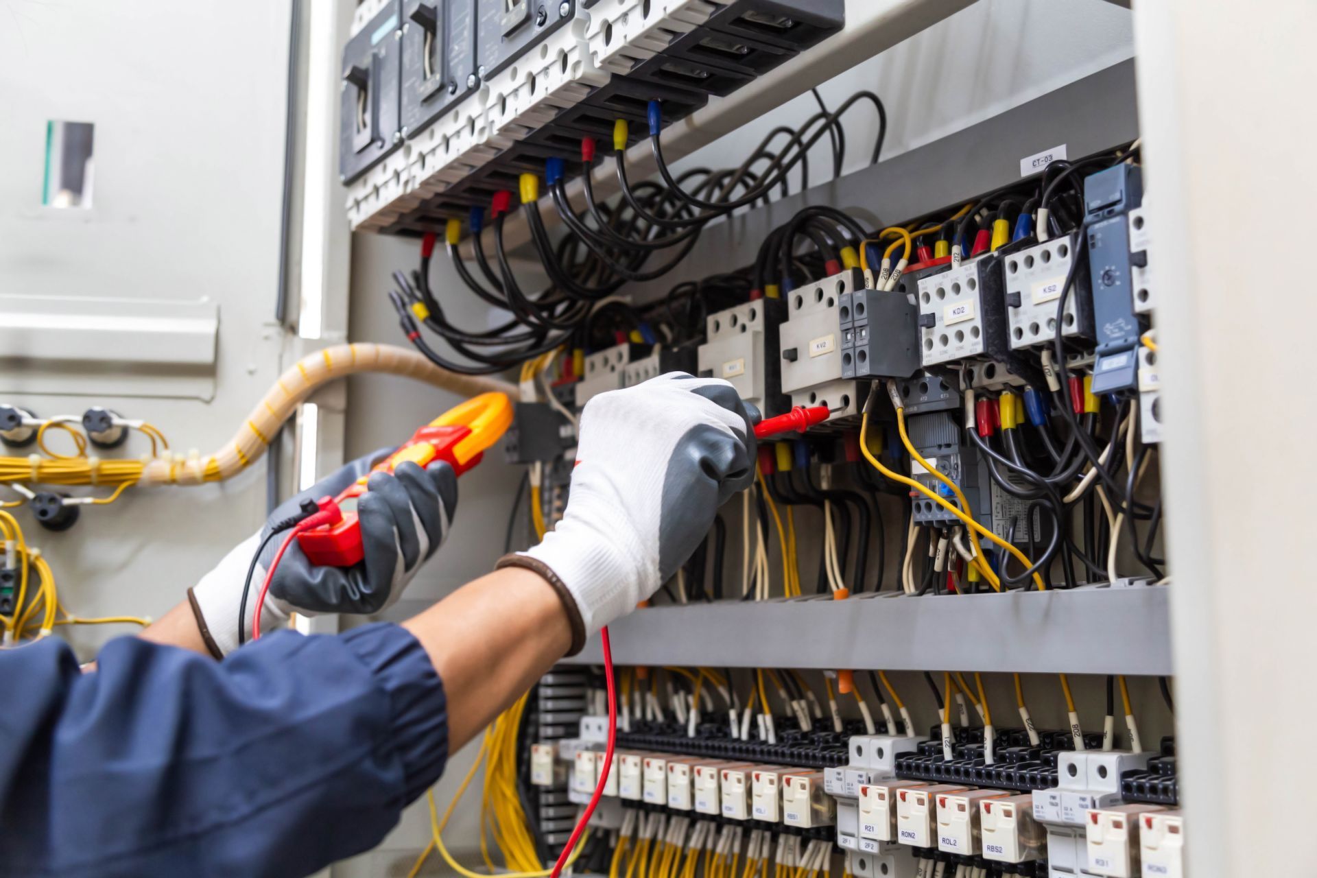 Electrician using a multimeter to inspect wiring in an electrical panel, wearing gloves.