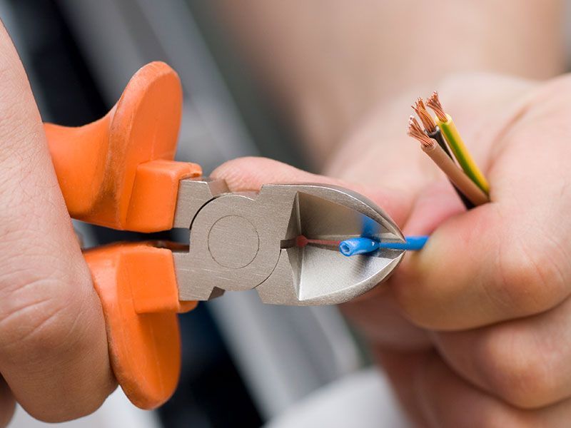 Hands using pliers to cut a blue electrical wire. Brown, yellow, and green wires are exposed.