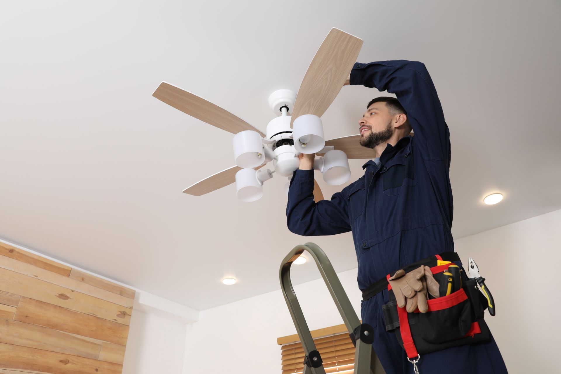Electrician on a ladder installing a ceiling fan with lights in a room.