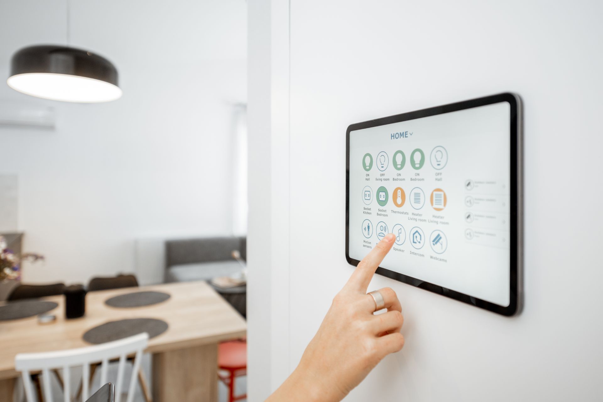 Person's finger touching a smart home control panel mounted on a white wall in a modern dining room.