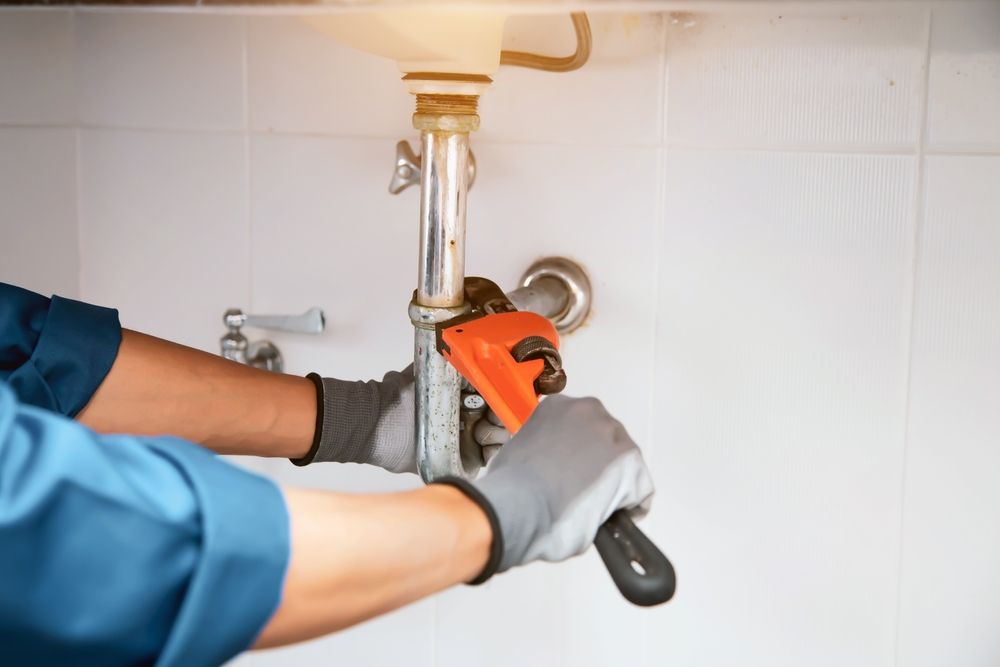 Plumber Using A Wrench On A Pipe Under A Sink, Wearing Work Gloves, In A Bathroom — Accredited Plumbing in Milton, NSW