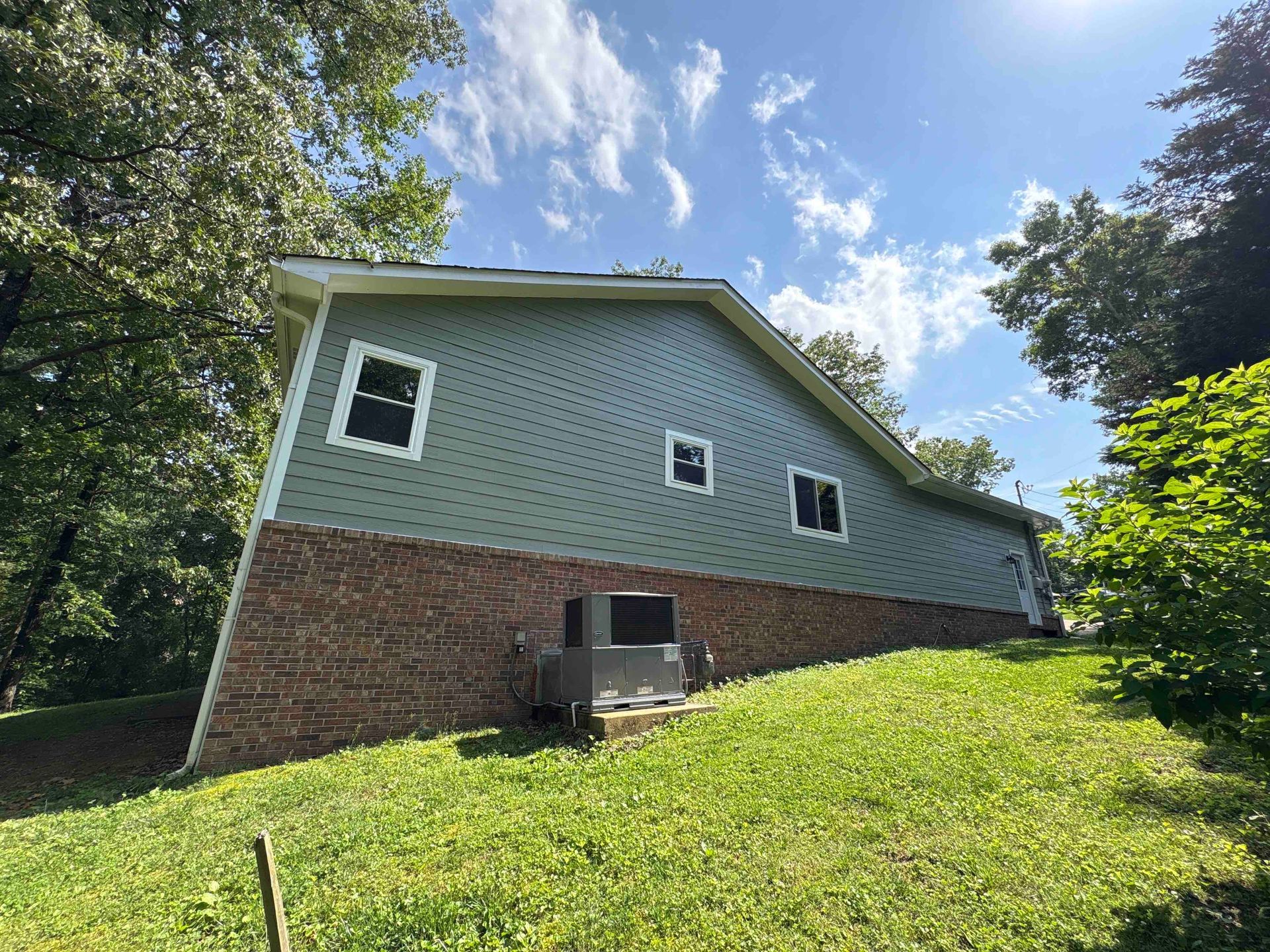 A house with a green siding is sitting on top of a grassy hill.