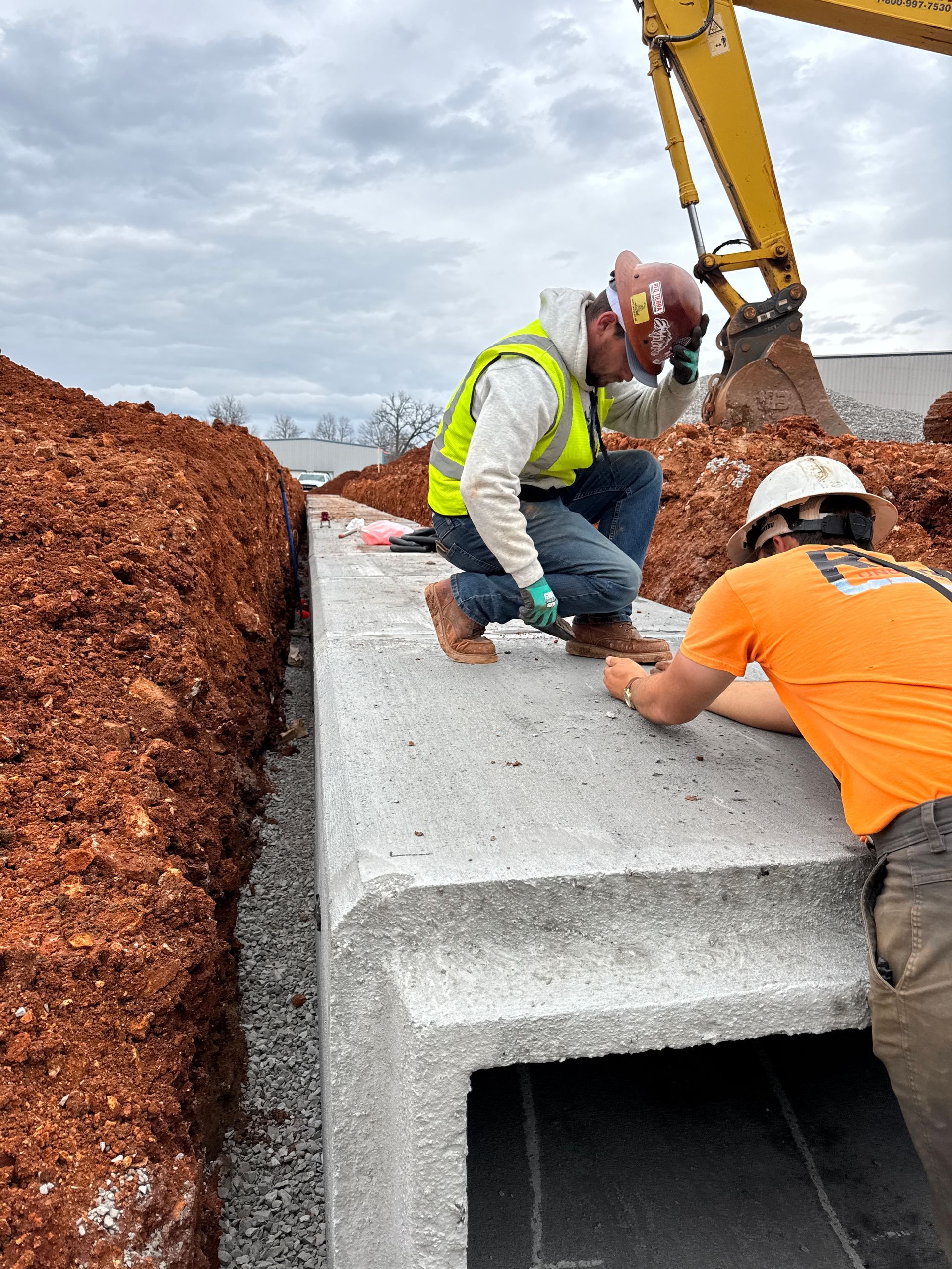 Two construction workers installing a concrete structure. One kneels, one stands; an excavator is in the background.
